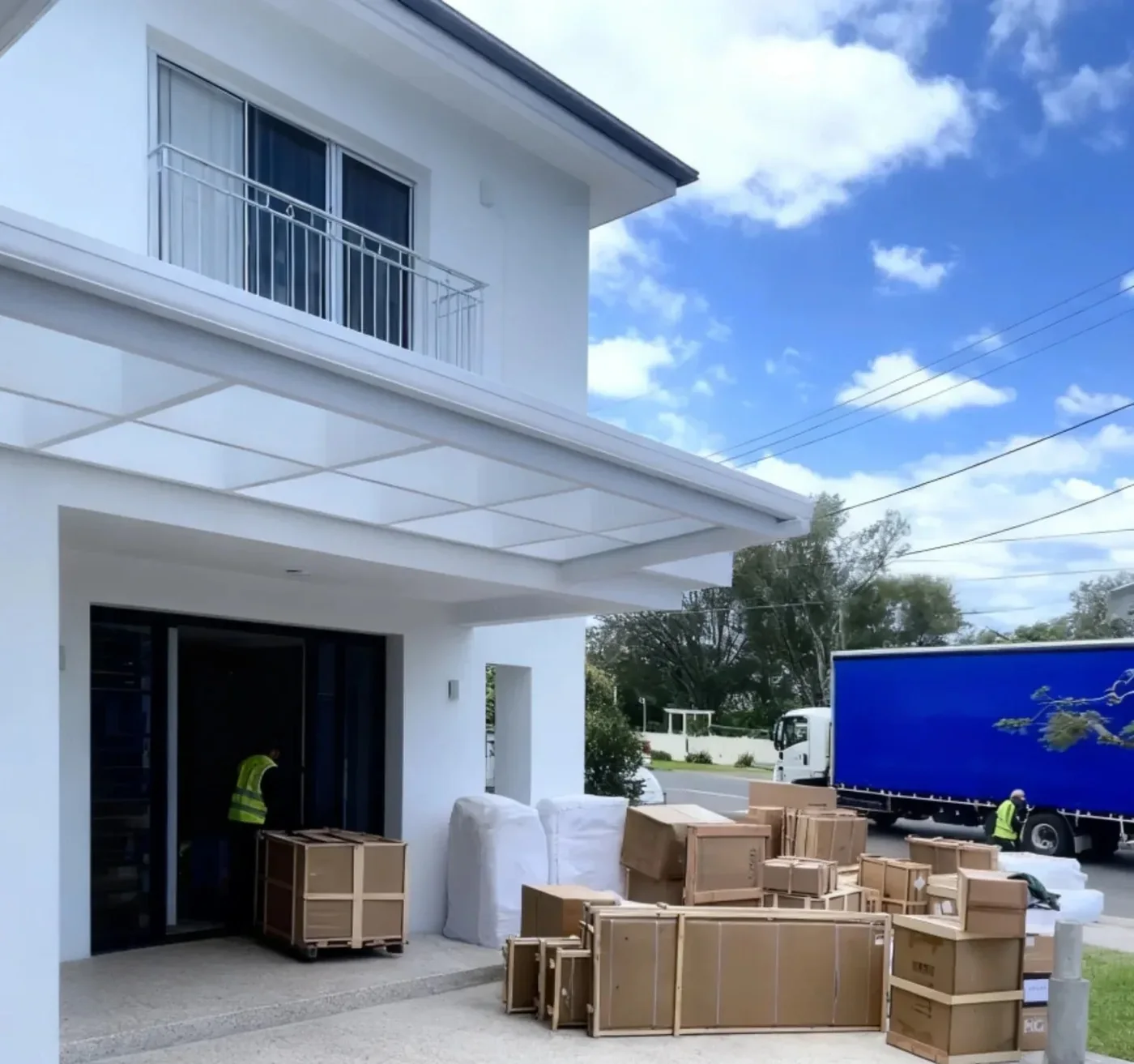 A curtain-side delivery truck parked on a US residential street with crates and wrapped furniture at the front door