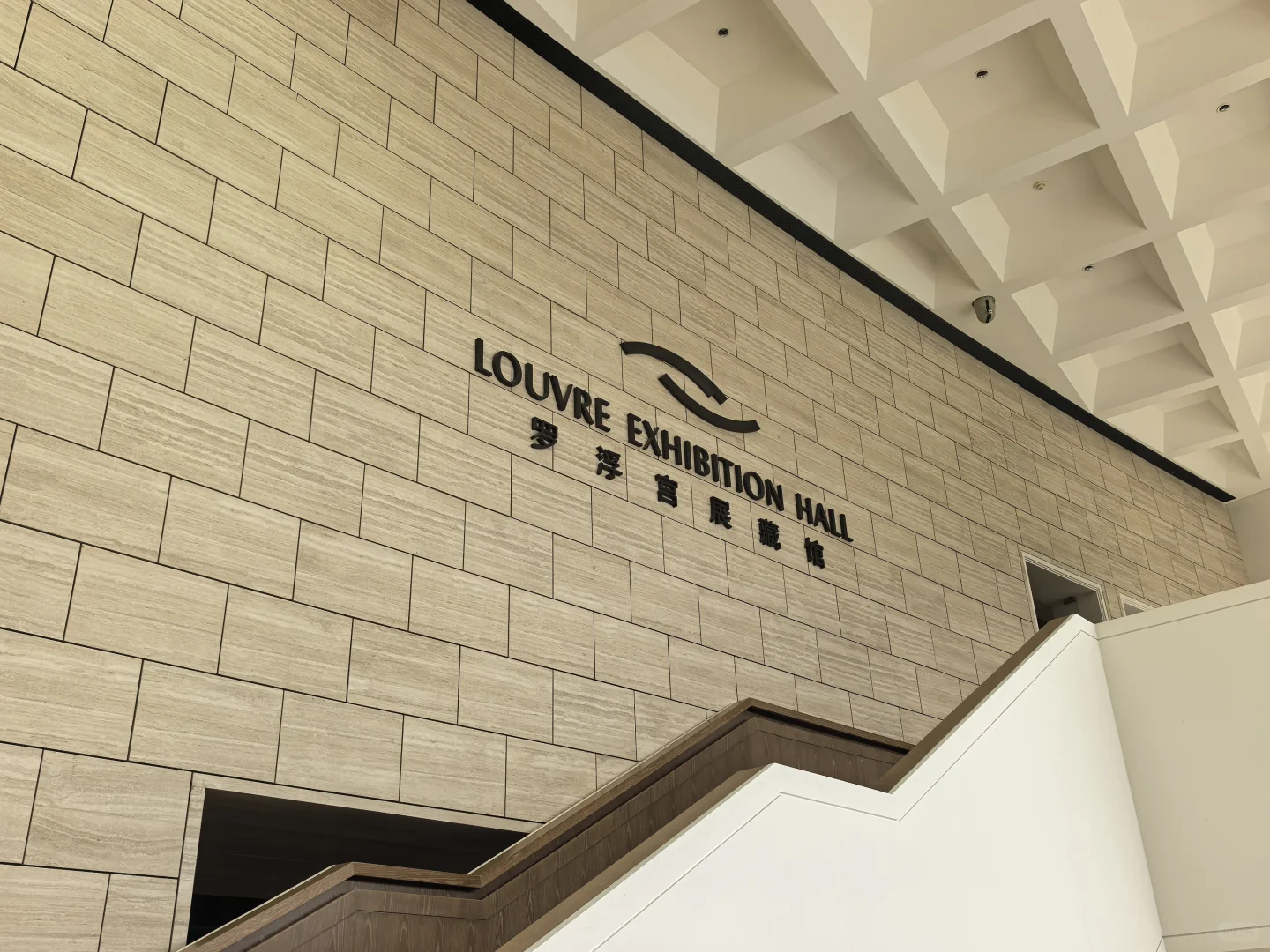 Louvre Exhibition Hall lettering on a cream stone wall above an angular staircase beneath a coffered ceiling.