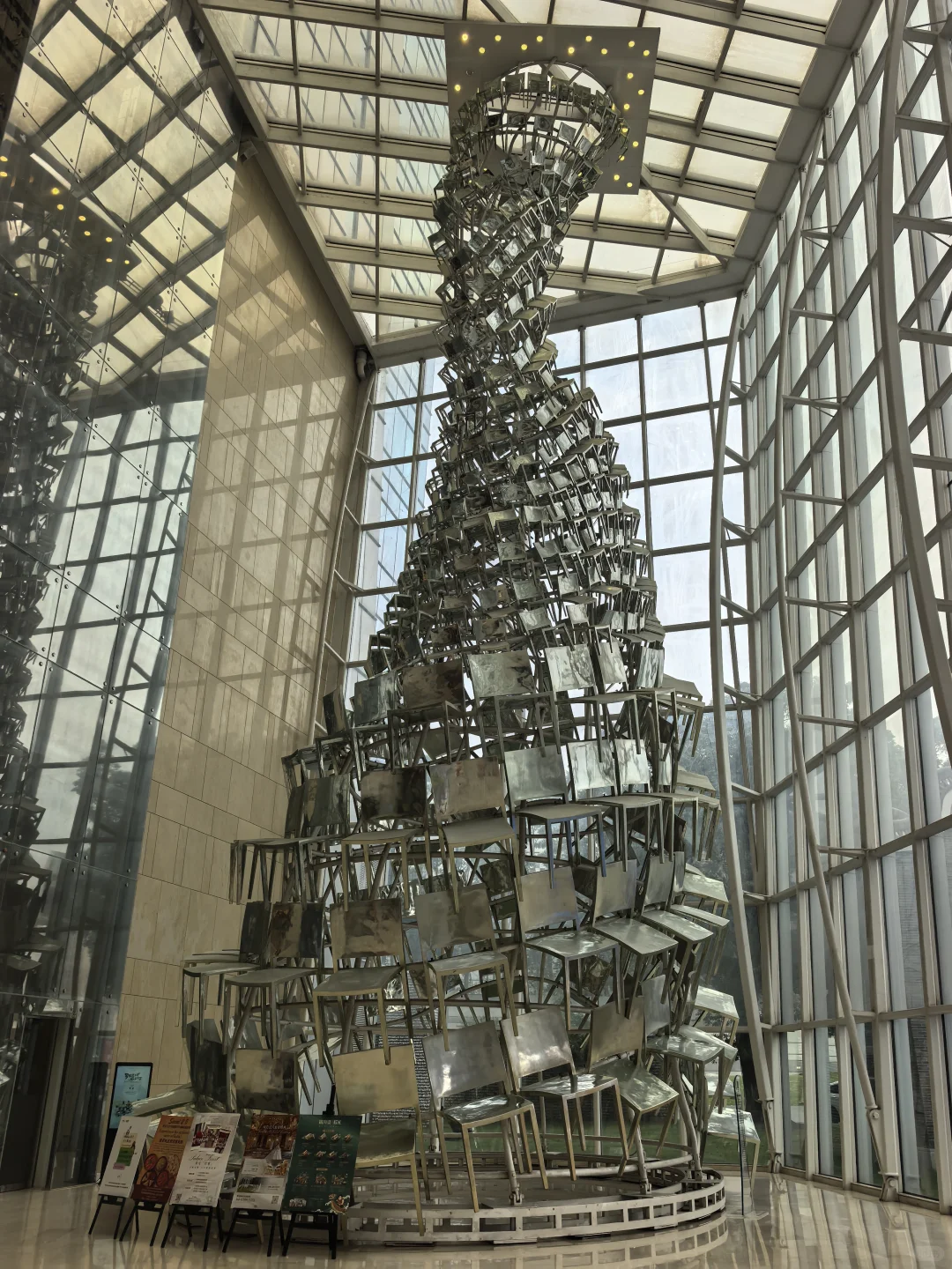 A twisting tower made from stacked metal chairs rises through a glass-roofed atrium, reflected in a mirrored wall.