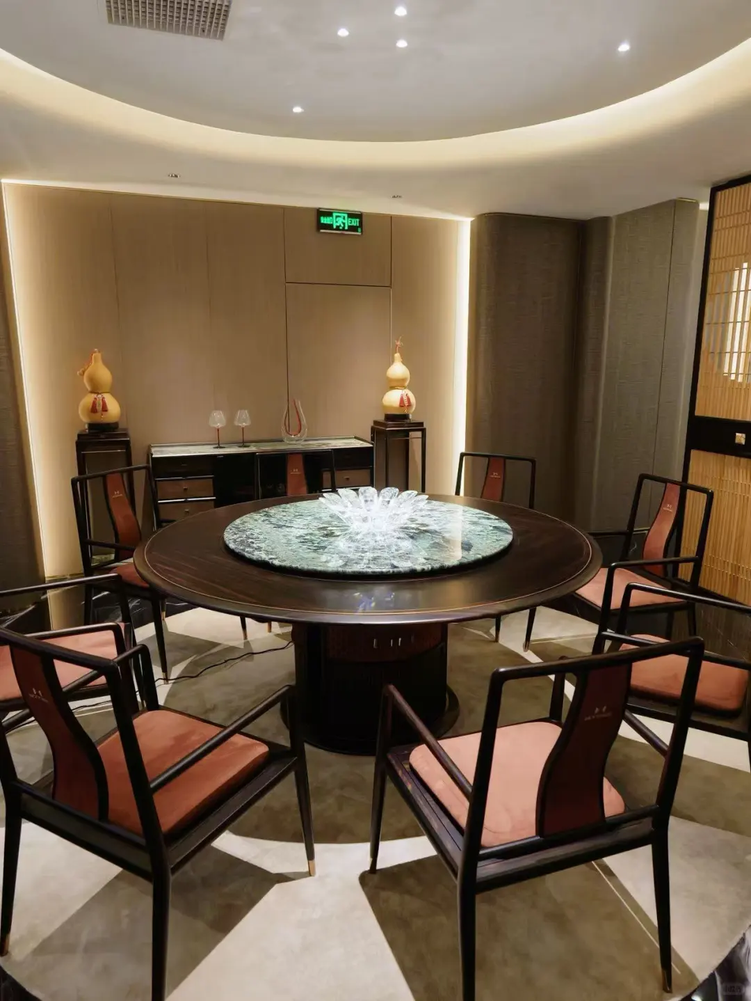 Round dark-framed dining table with a green-toned centre and red upholstered chairs in a softly lit room.