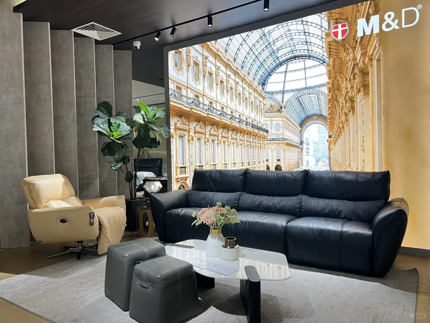 Black M&D sofa with padded back cushions, grey stools and a light-coloured coffee table in a display setting.
