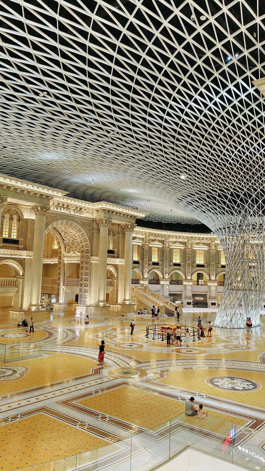 Wide view across a patterned plaza toward classical arches beneath branching lattice supports.