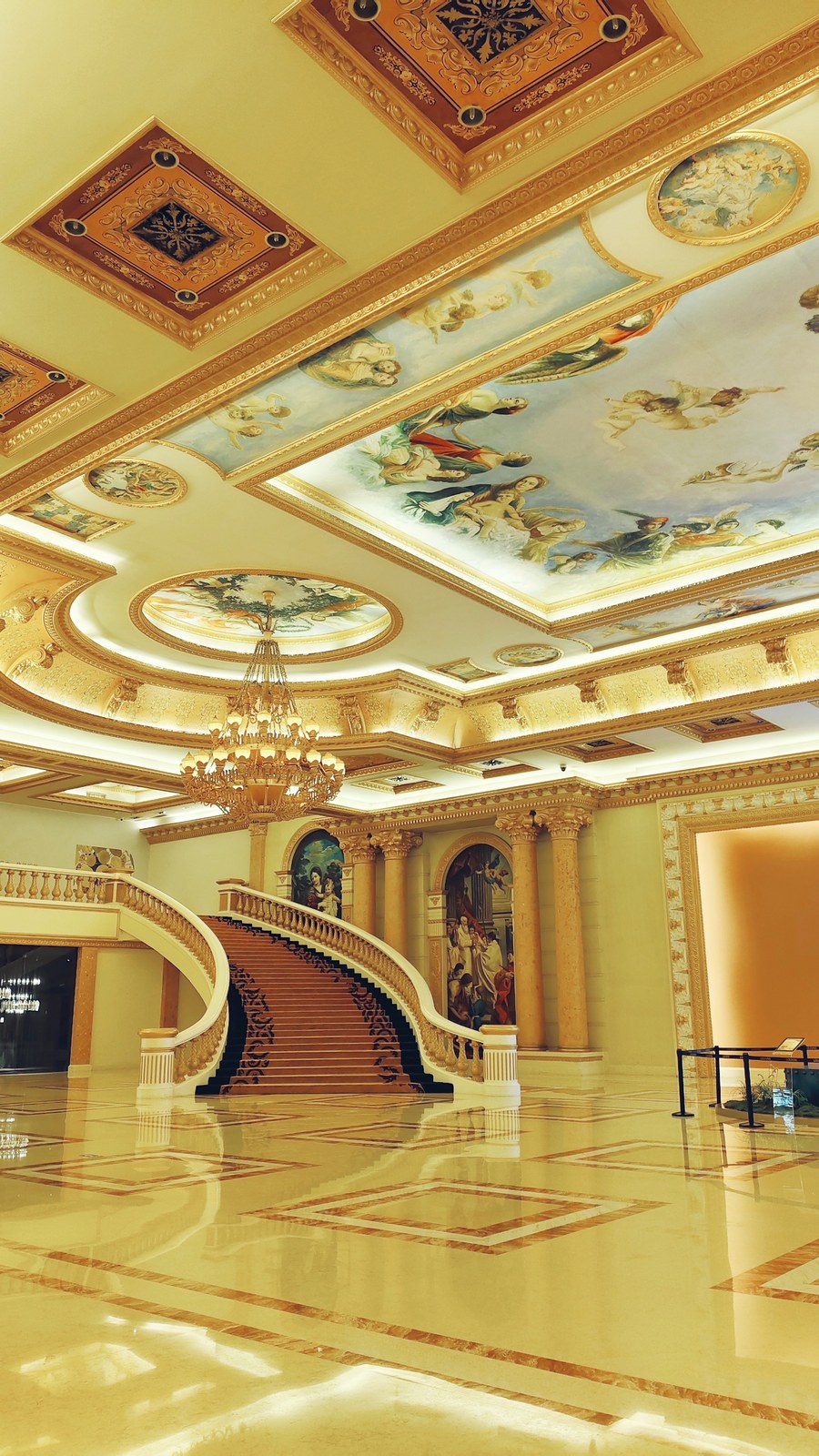 Curving staircase beneath a large chandelier in a gold-toned hall with painted ceiling panels.