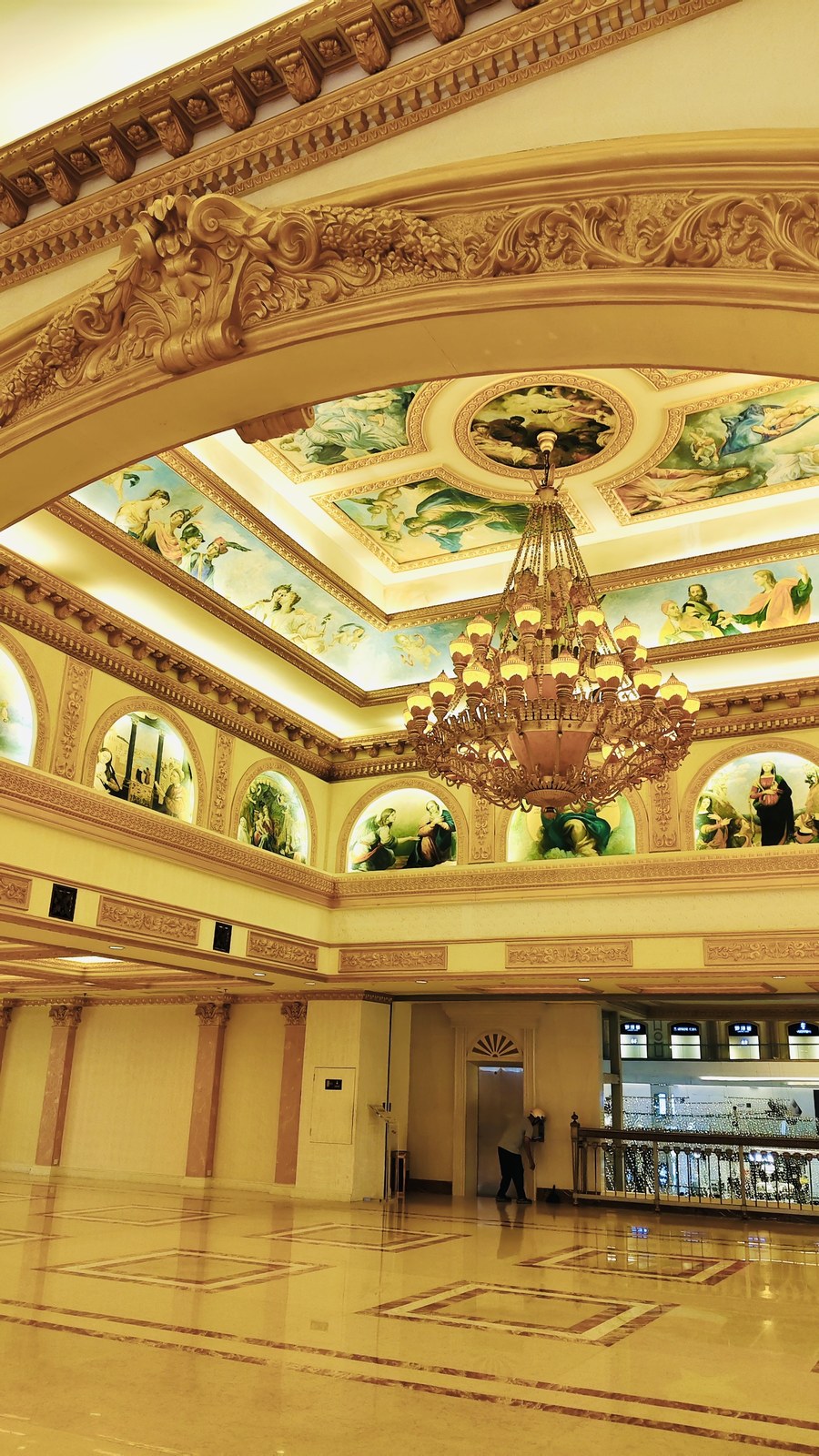 Multi-tier chandelier surrounded by painted ceiling panels and elaborate gold-toned mouldings.