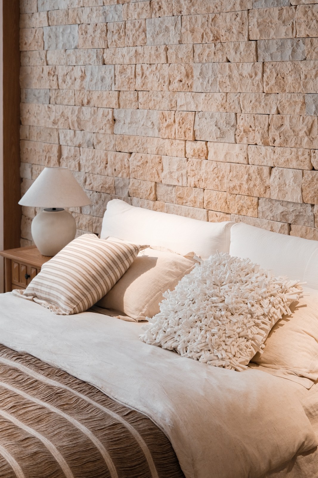 Close view of neutral bedding, striped and textured cushions, and a pale bedside lamp.