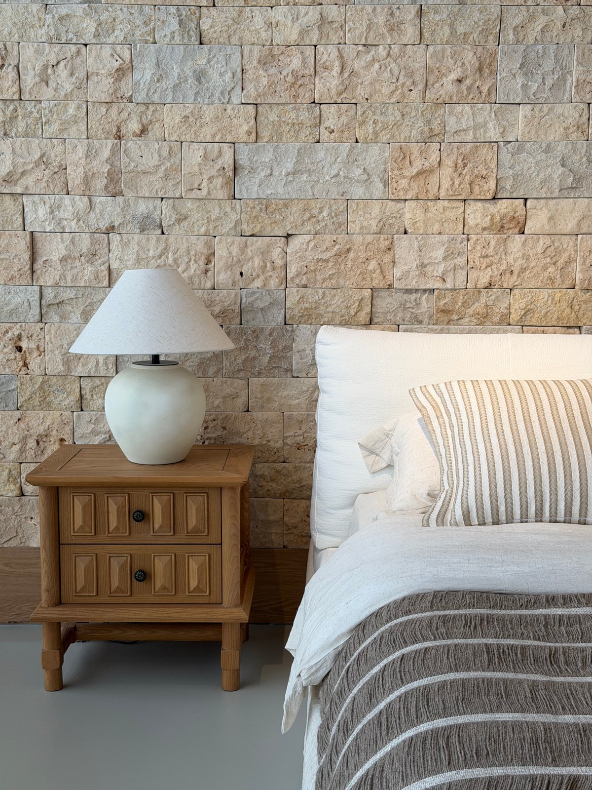 Two-drawer wood-toned bedside cabinet and pale lamp next to neutral bedding.