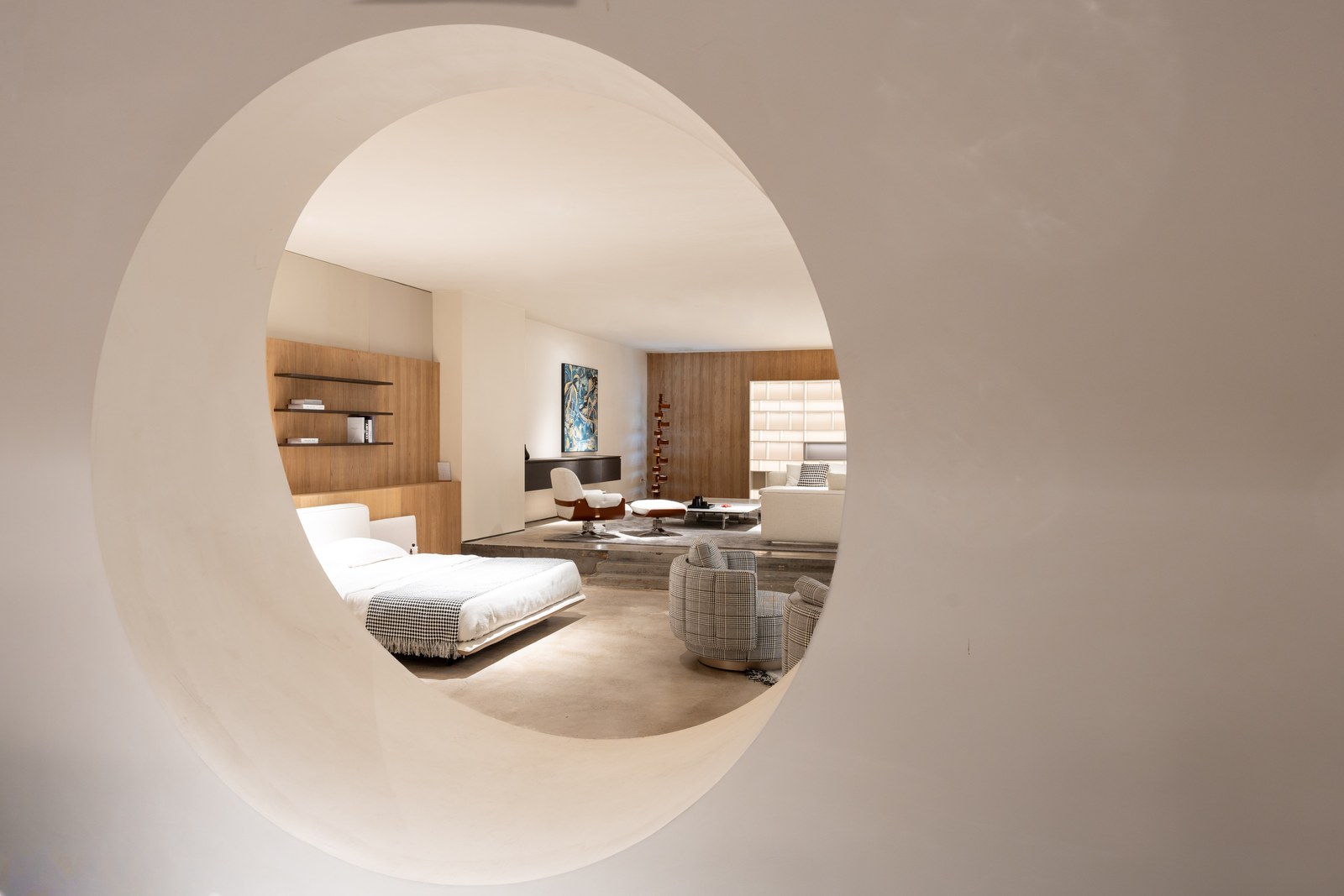 Round architectural opening overlooking a white bed, patterned chairs and a raised sitting area.