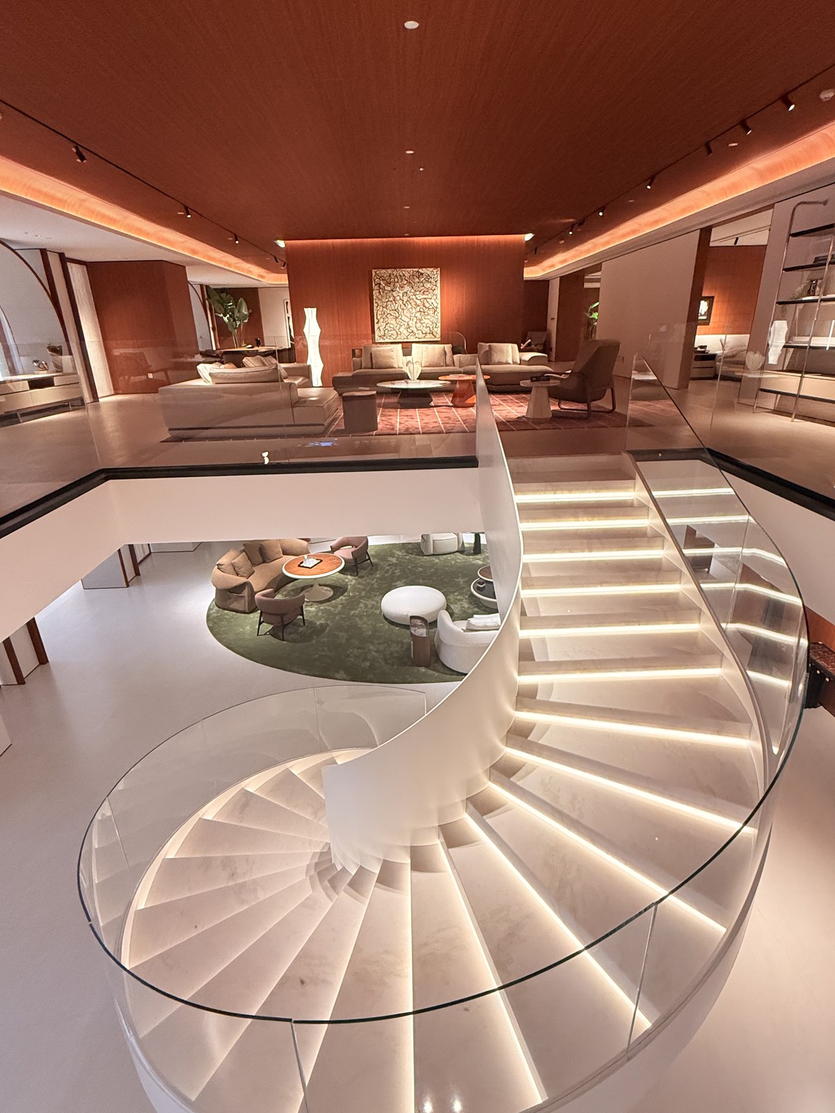 Curved staircase with illuminated treads and glass balustrades between two furniture-display floors.