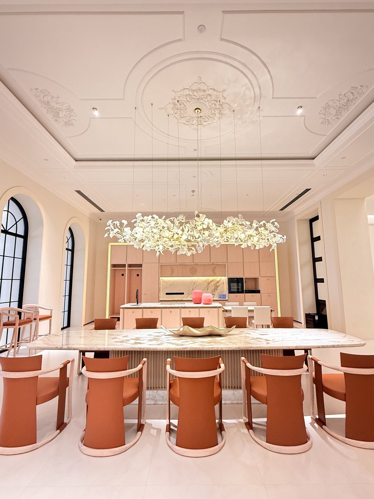 Long pale veined dining table with terracotta-toned chairs beneath a leafy pendant installation.