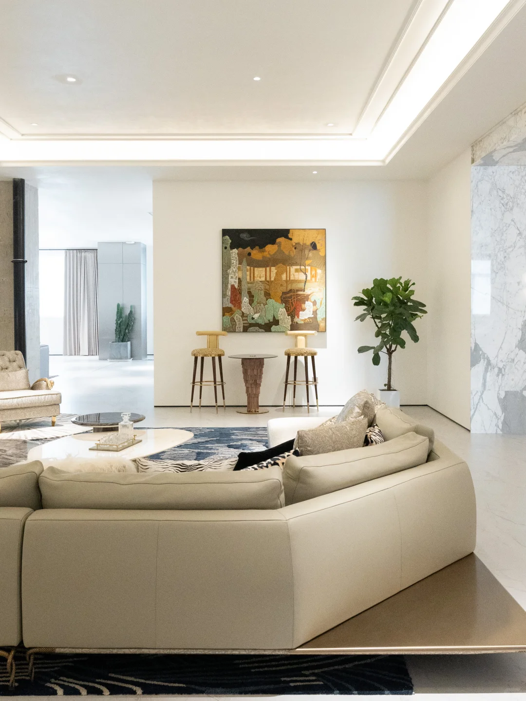 Cream sectional sofa viewed from the rear with layered coffee tables, a blue patterned rug, two tall stools and abstract artwork behind it.