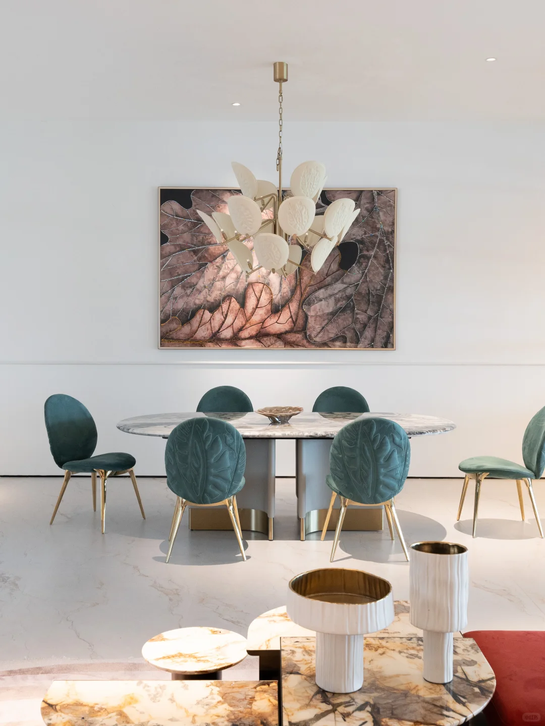 Six teal upholstered dining chairs with leaf-patterned backs around an oval grey-veined table beneath a white leaf-shaped chandelier.