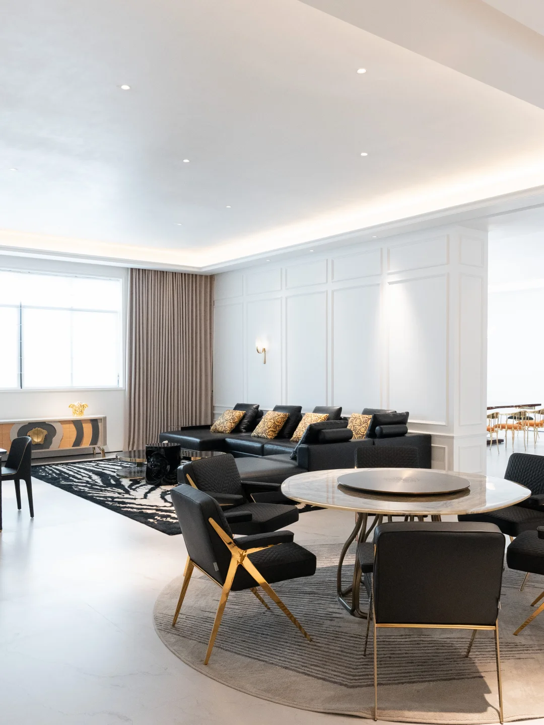 Round light-coloured dining table and black chairs with gold-toned legs in front of a black sectional sofa and patterned rug.