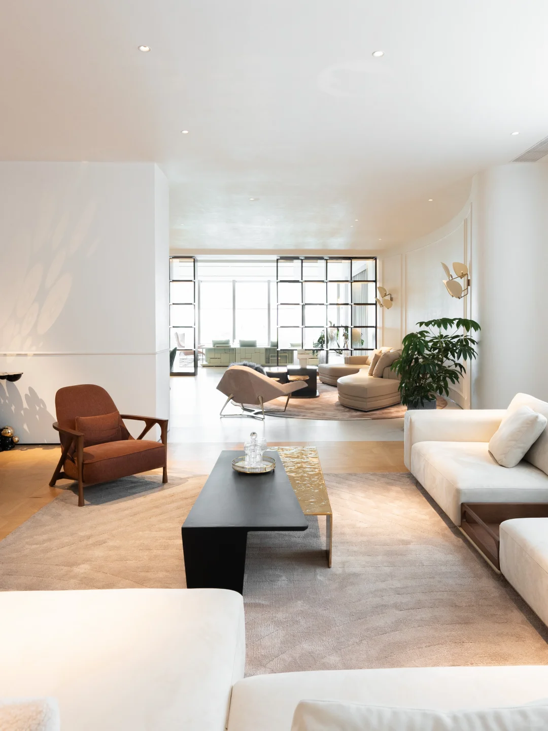 Brown armchair and white sofa facing a long black coffee table, with a second curved seating group beside bright windows behind them.