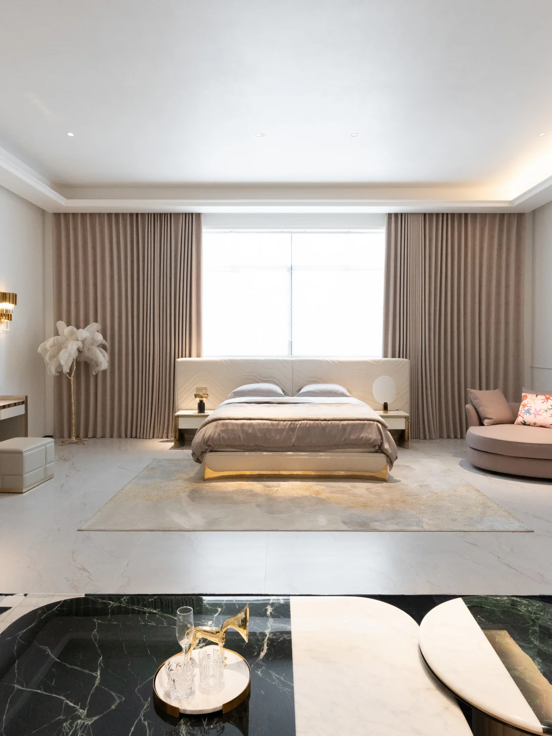 Cream upholstered bed centred beneath a bright window, with full-height taupe curtains, paired bedside tables and a curved chair at the right.