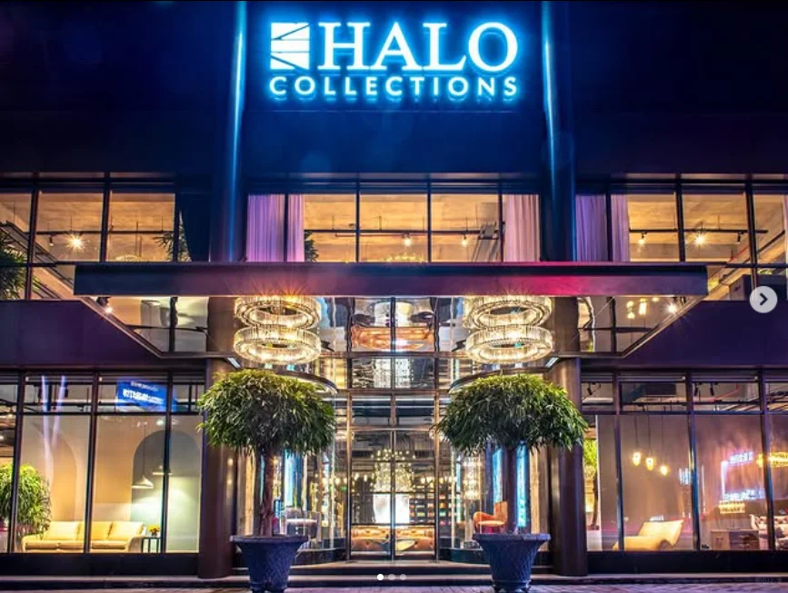 Glass-fronted Halo Collections entrance with a bright blue brand sign, two round-canopy trees and chandelier lighting beneath a mirrored canopy.