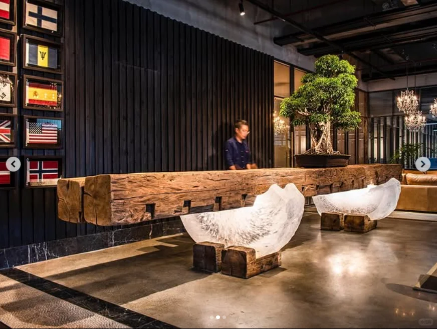 Long rough-textured reception counter supported by two translucent curved forms, with a potted tree, a dark ribbed wall and framed flags behind it.