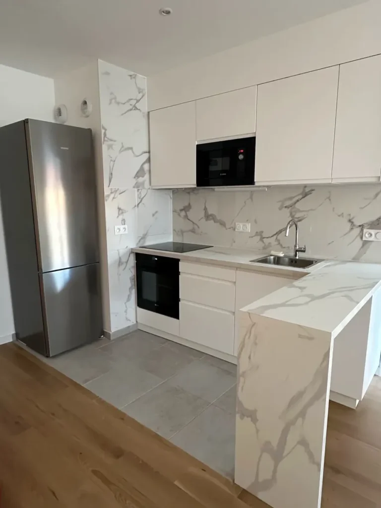 A finished small kitchen in a Paris new-build: matt white handle-less units, full-height stone-effect splashback and an L-shaped breakfast bar.
