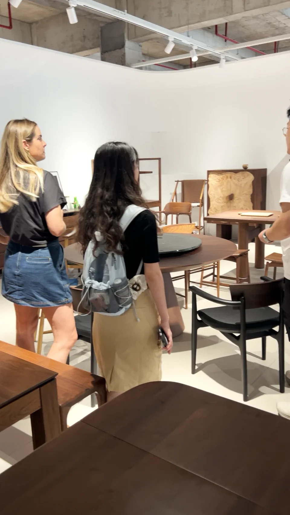 A buyer and guide walking among timber tables and chairs in a furniture showroom.